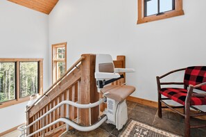 A wooden staircase with a handrail, a chair with a checkered cushion, and a window with a view of trees.