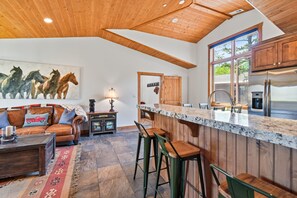 A spacious kitchen with a wooden ceiling, a large island with bar stools, and a dining area with a leather couch and a rug.