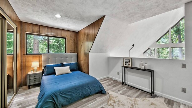 Un dormitorio con techo inclinado, una cama con ropa de cama azul, una mesita de noche, una lámpara de noche, un cuadro enmarcado y una consola con botellas.