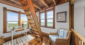 A room with a wooden ladder, a wicker chair, a desk with a lamp, and large windows with a view. - The Gothic House with Beachfront (Pensacola)
