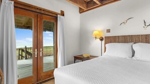 Quarto de hotel com uma cama, mesa de cabeceira, luminária e vista para a praia através de uma porta de vidro.