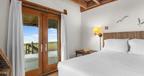 Ein Hotelzimmer mit Bett, Nachttisch, Lampe und Blick auf den Strand durch eine Glastür.