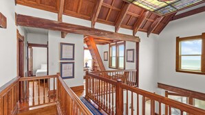 Uma escadaria de madeira com corrimão, que leva a um quarto com cama e janela com vista para o mar.