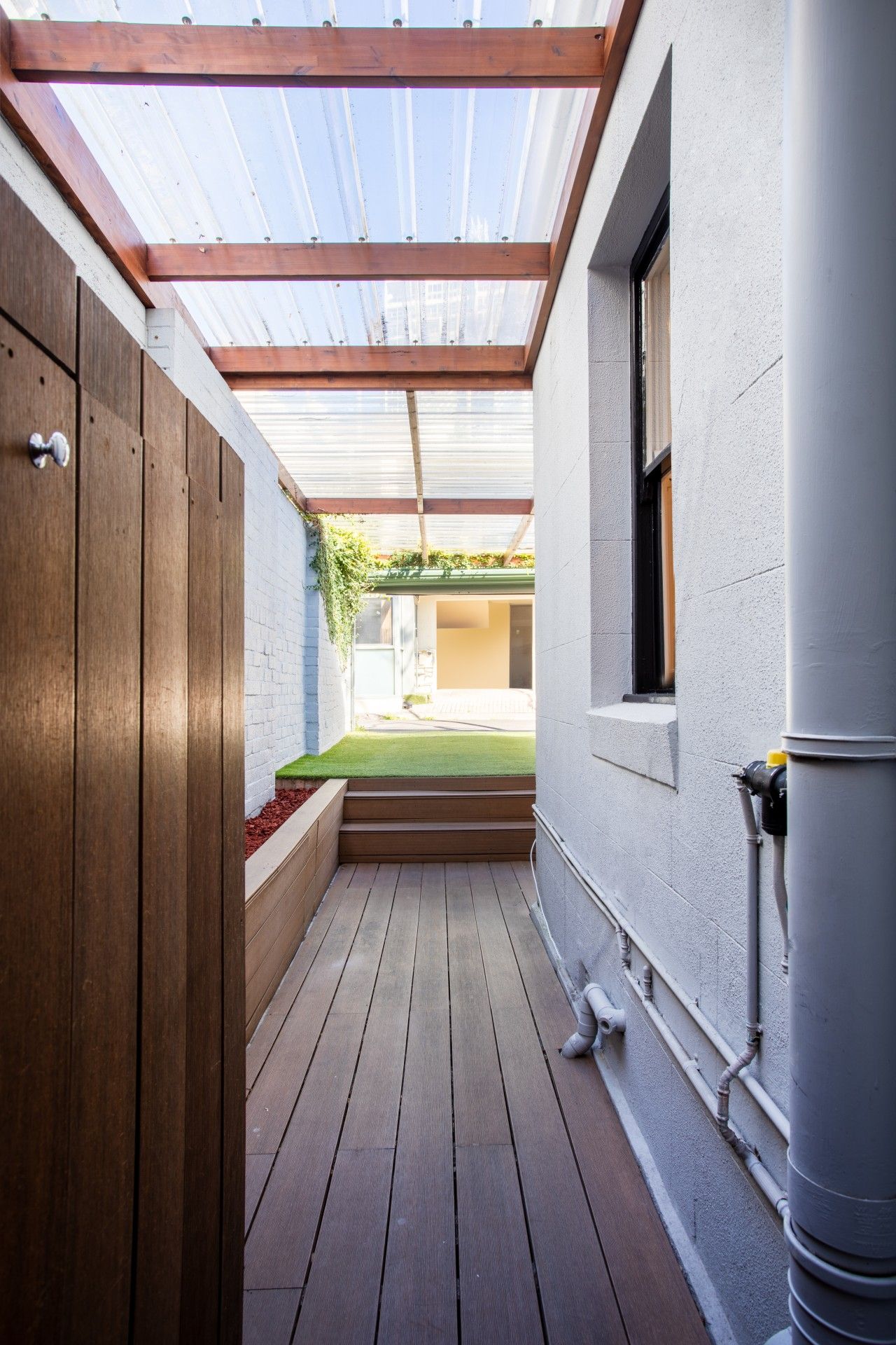 Un espace extérieur abrité doté d’un plancher en bois, d’une fenêtre et d’un mur blanc avec des tuyaux apparents.