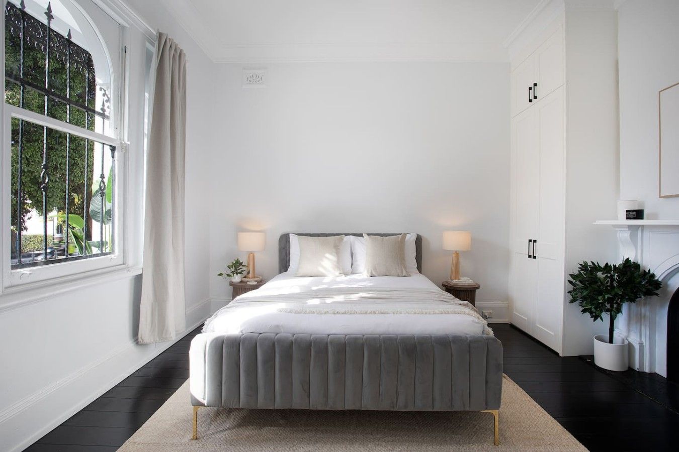 A bedroom with a large bed, two bedside tables, a window with curtains, a fireplace, and a potted plant.
