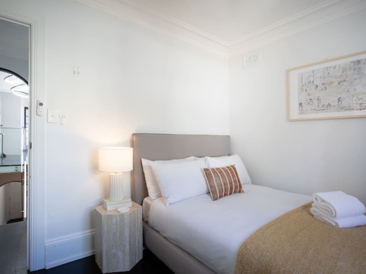 Une chambre d’hôtel avec un lit, une table de chevet avec une lampe, une œuvre d’art encadrée au mur et une porte donnant sur une autre pièce.