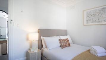 Une chambre d’hôtel avec un lit, une table de chevet avec une lampe, une œuvre d’art encadrée au mur et une porte donnant sur une autre pièce.
