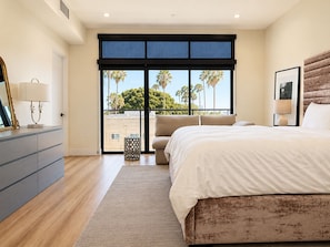 Ein Schlafzimmer mit einem großen Bett, einer Kommode, einem Fenster mit Blick auf Palmen und einer Decke mit Einbaustrahlern.
