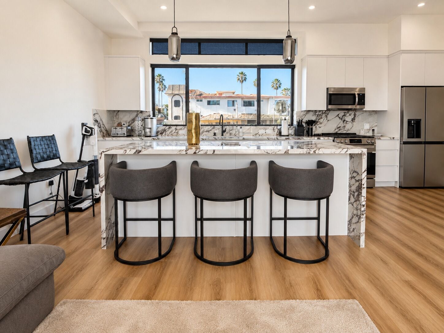A modern kitchen with a marble countertop, stainless steel appliances, and a large window with a view of palm trees.