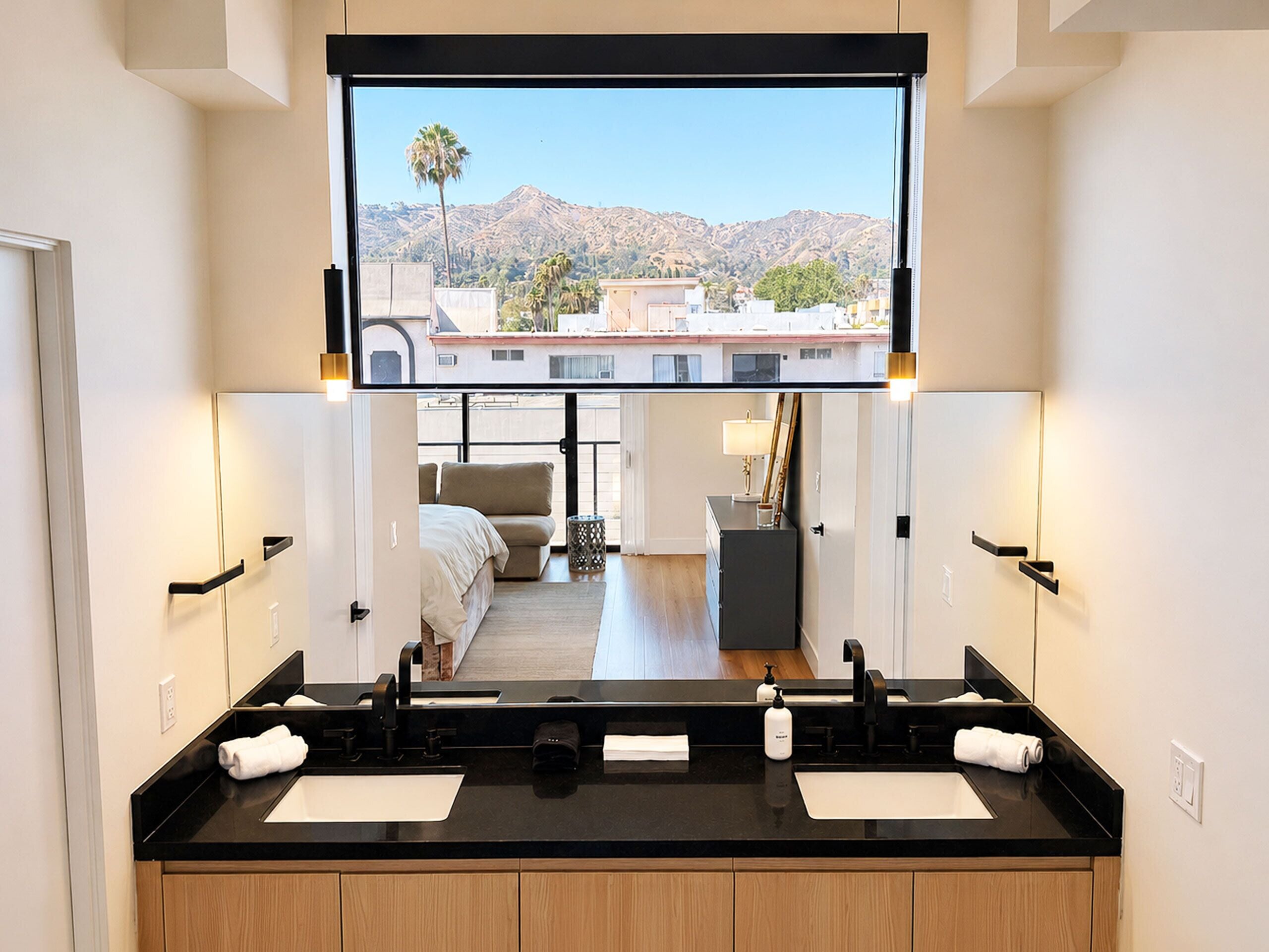 Un baño moderno con encimera negra, dos lavamanos y un espejo grande que refleja un dormitorio y vistas de montañas.