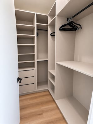 Ein begehbarer Kleiderschrank mit mehreren Regalen und Ablagefläche zum Aufhängen, mit Holzboden und einer Tür mit schwarzem Griff.