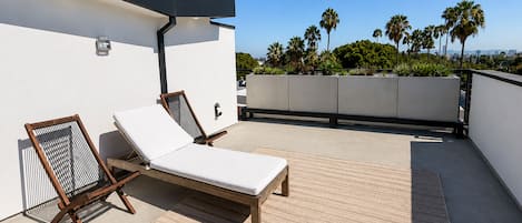 A rooftop terrace with two sun loungers, a wall-mounted light fixture, and a view of palm trees and buildings in the distance.