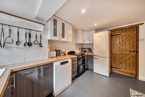 A compact kitchen with white cabinets, a dishwasher, a stove, and a refrigerator. There is a wooden door and a hanging utensil rack. - Quaint Cottage in Charming Evesham (Evesham)