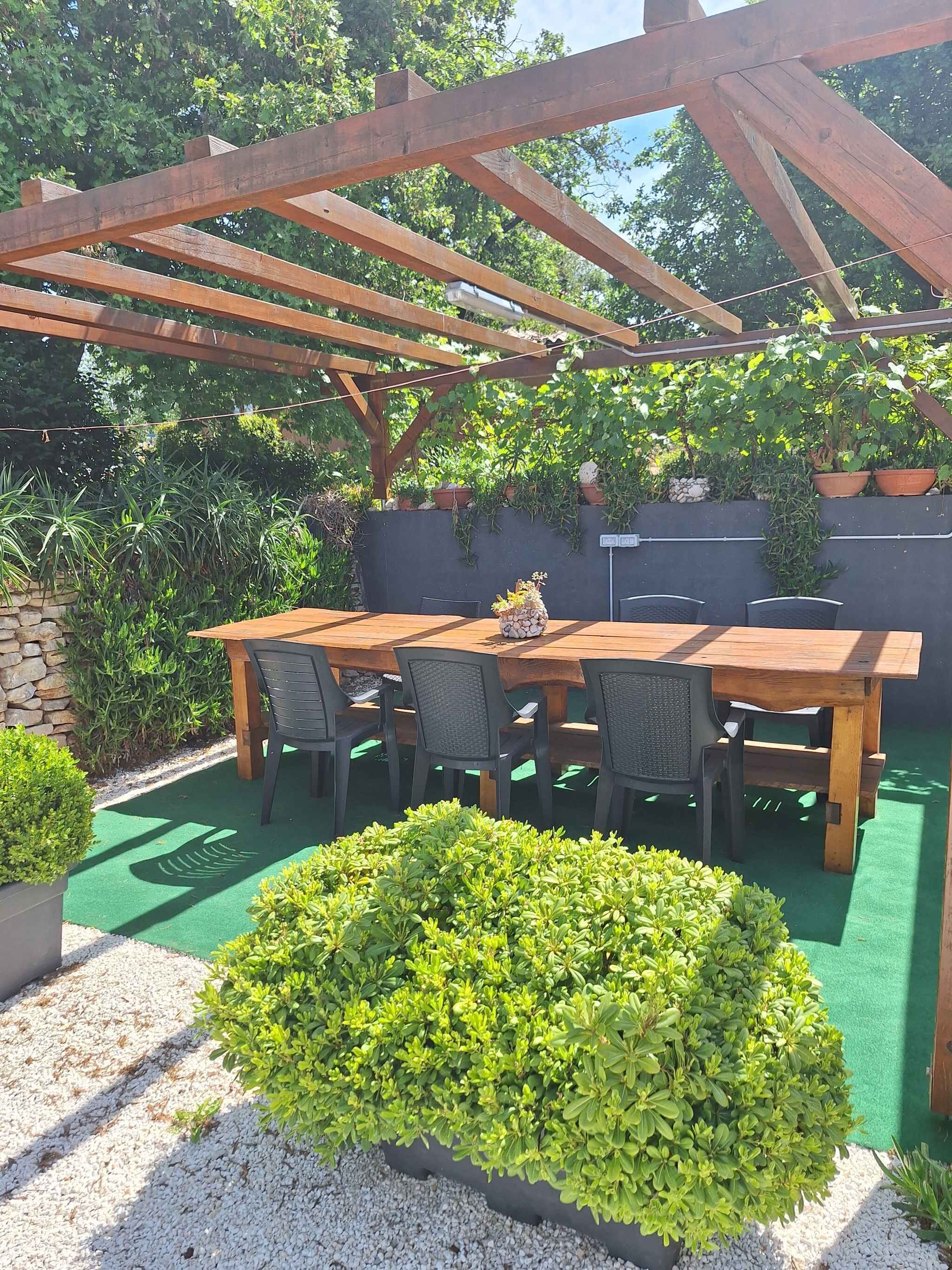 A wooden pergola with a table and chairs, surrounded by greenery and a gravel path.