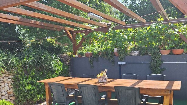 En træpergola med et bord og stole, omgivet af grønt og en grussti.