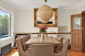 A dining area with a large table, six chairs, a chandelier, a mirror, and a vase with flowers.