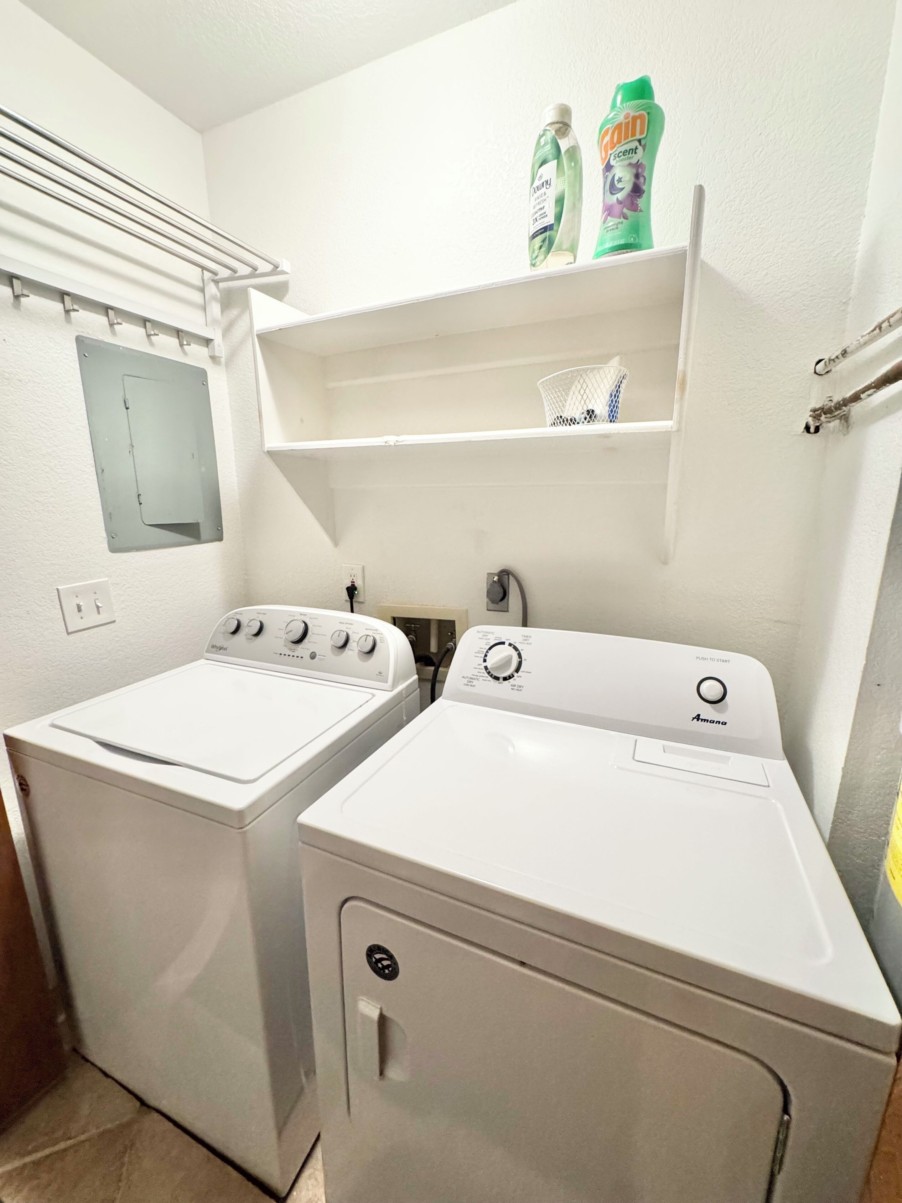 A laundry room with two white washing machines, a metal drying rack, and a shelf with cleaning products.
