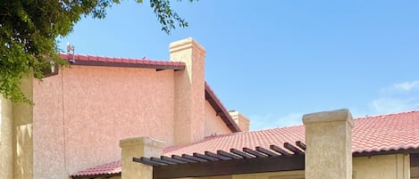 A residential building with a red-tiled roof, a covered patio area, and a small garden with a tree and flowering plants.