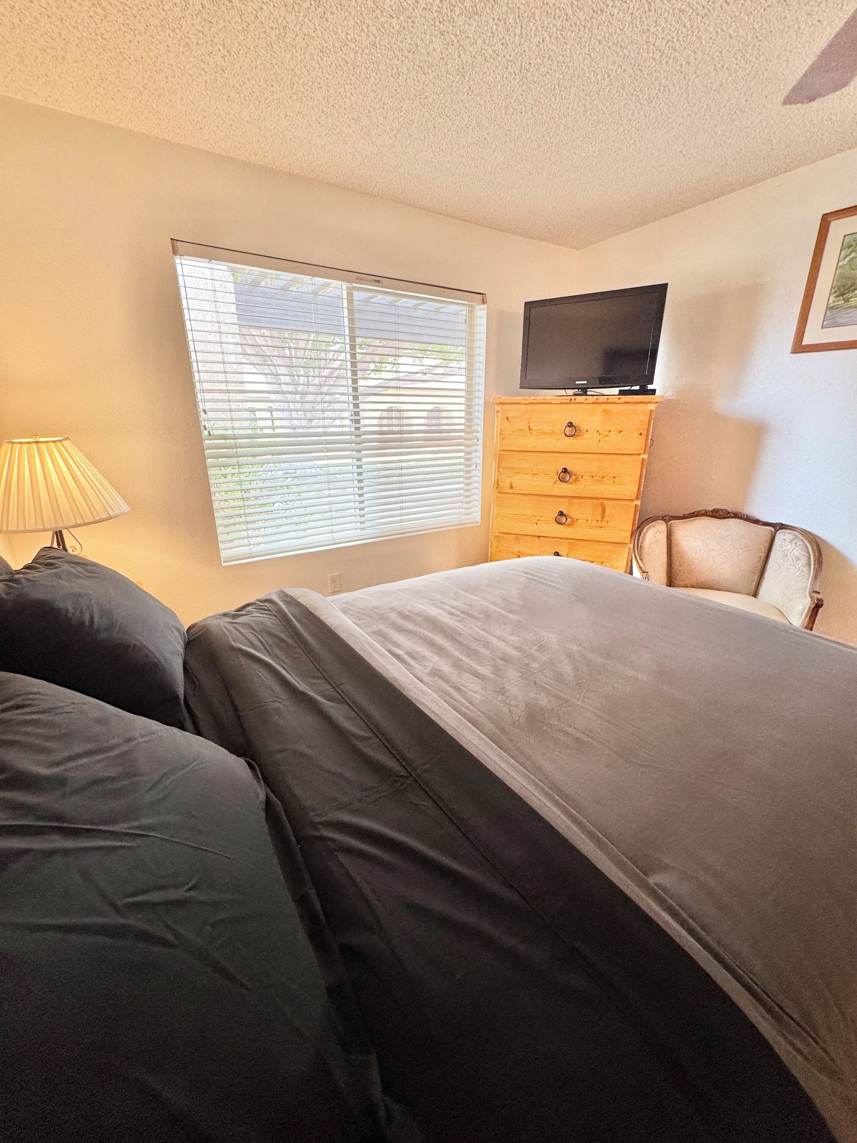 A bedroom with a bed, a dresser, a television, and a window with blinds.