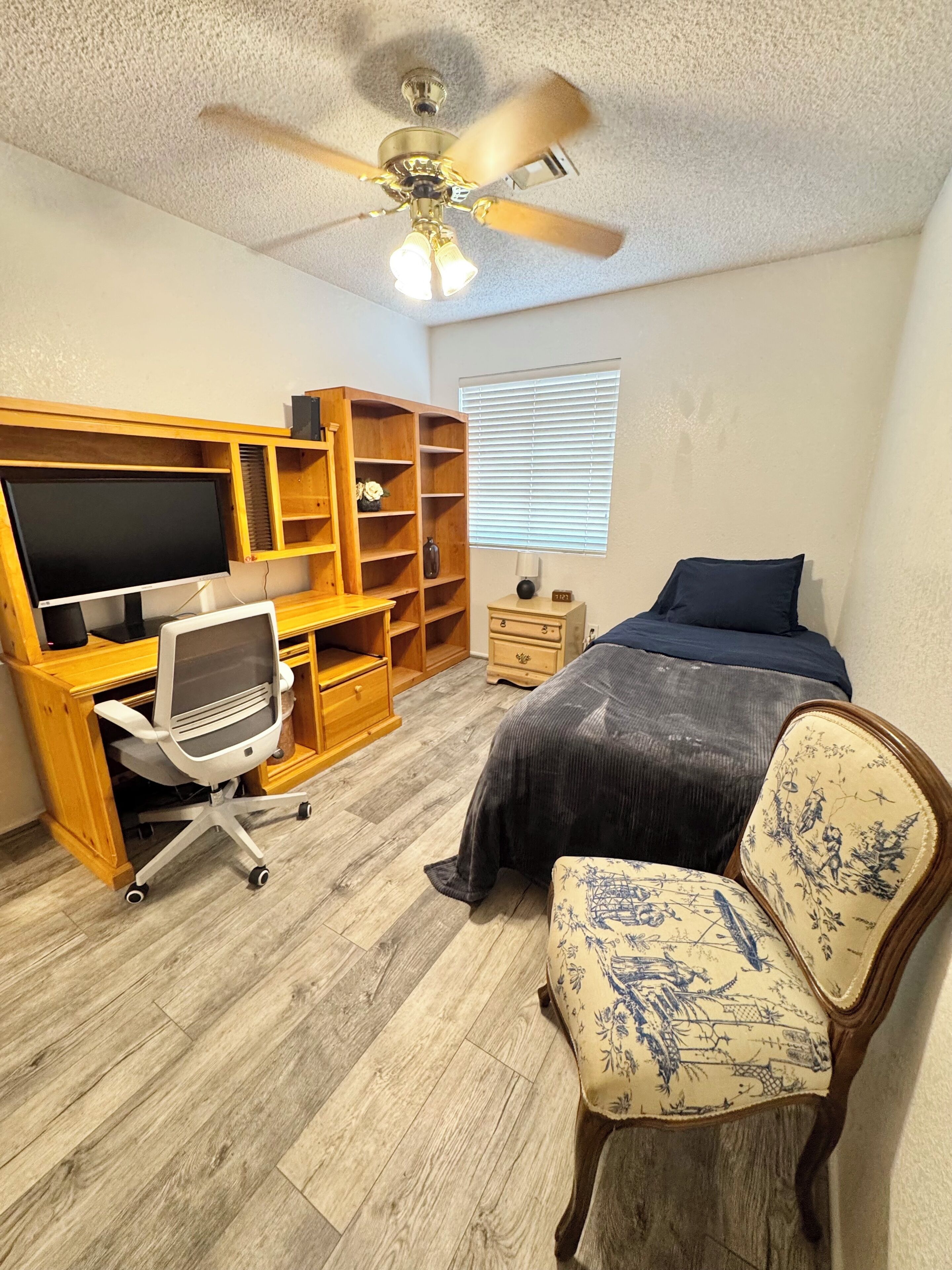 A bedroom with a bed, a desk with a computer, a chair, a ceiling fan, and a window.