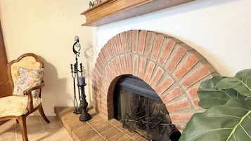 A fireplace with a brick surround, a mantel shelf with decorative items, a chair with a cushion, and a large green plant.