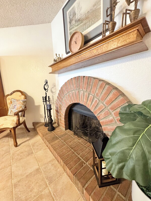A fireplace with a brick surround, a mantel shelf with decorative items, a chair with a cushion, and a large green plant.