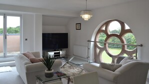 Ein modernes Wohnzimmer mit Fernseher, bequemen Sitzmöglichkeiten, einem Glastisch und einem großen Bogenfenster mit Blick auf Grünflächen.