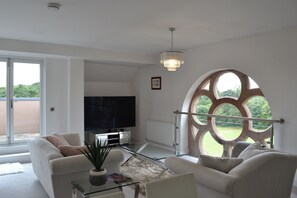 Een moderne woonkamer met een flatscreen televisie, comfortabele zithoek, een glazen salontafel en een groot, rondboogvenster met uitzicht op groen.
