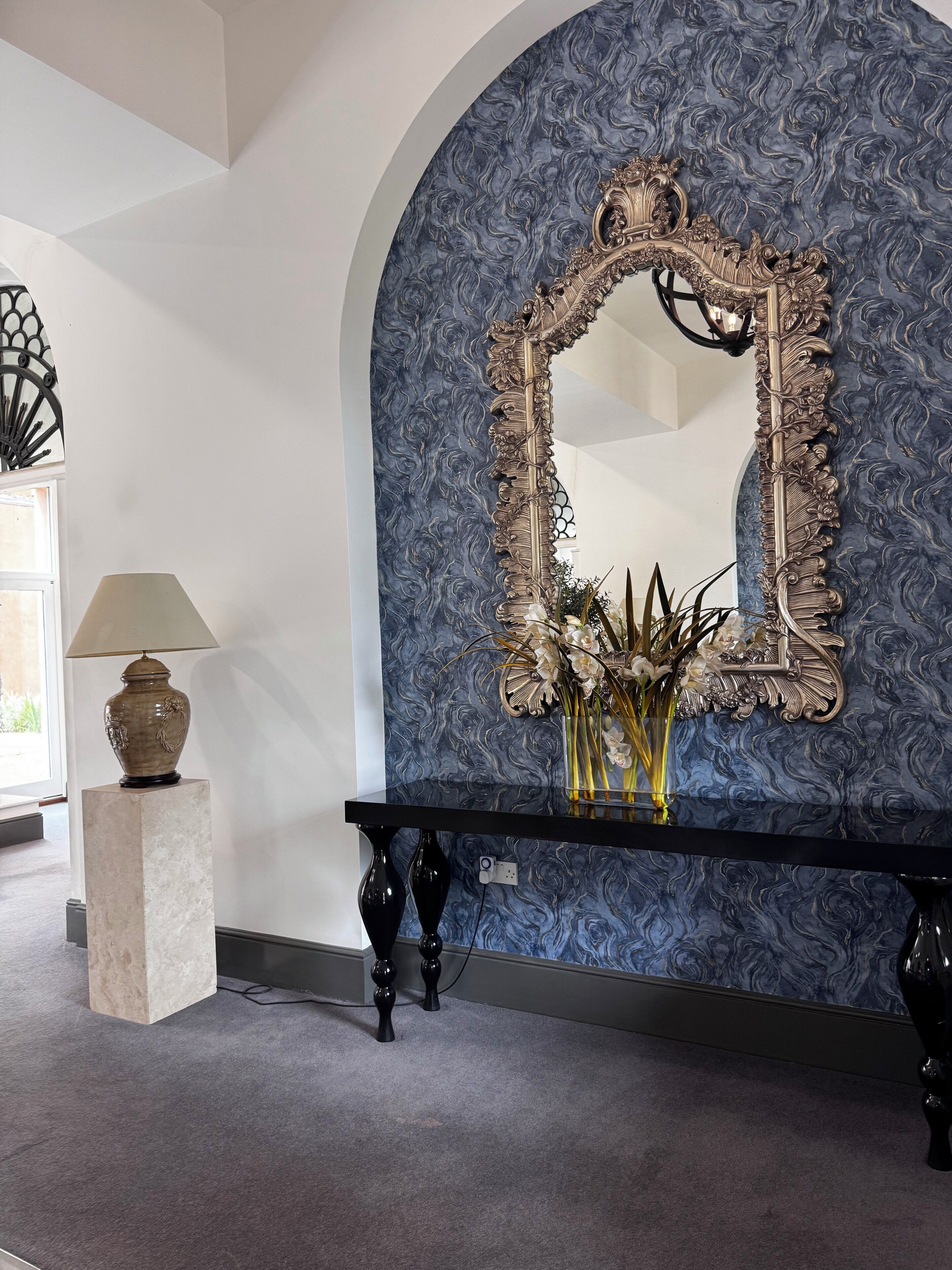 A room with a large ornate mirror, a console table with a vase of flowers, and a lamp on a pedestal.