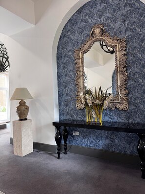 A room with a large ornate mirror, a console table with a vase of flowers, and a lamp on a pedestal.