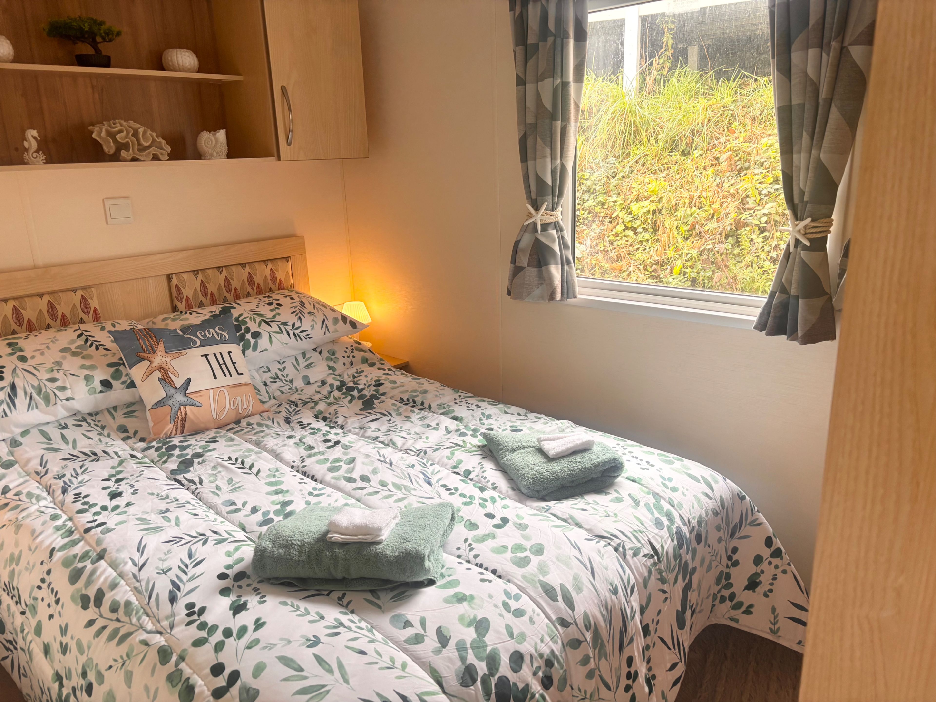 A bedroom with a bed, a nightstand, a window with curtains, and a shelf with decorative items.