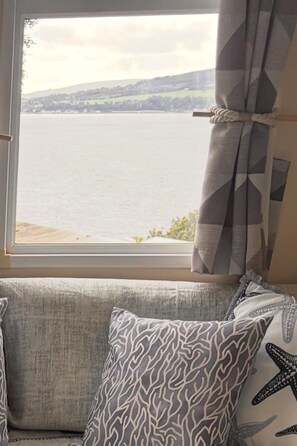 Ein Fenster mit Blick auf ein Gewässer und Hügel, ein Vorhang und ein Sofa mit gemusterten Kissen.