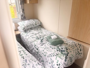 A small, single-bed room with a window, a bedside table with a lamp, and a green towel on the bed.