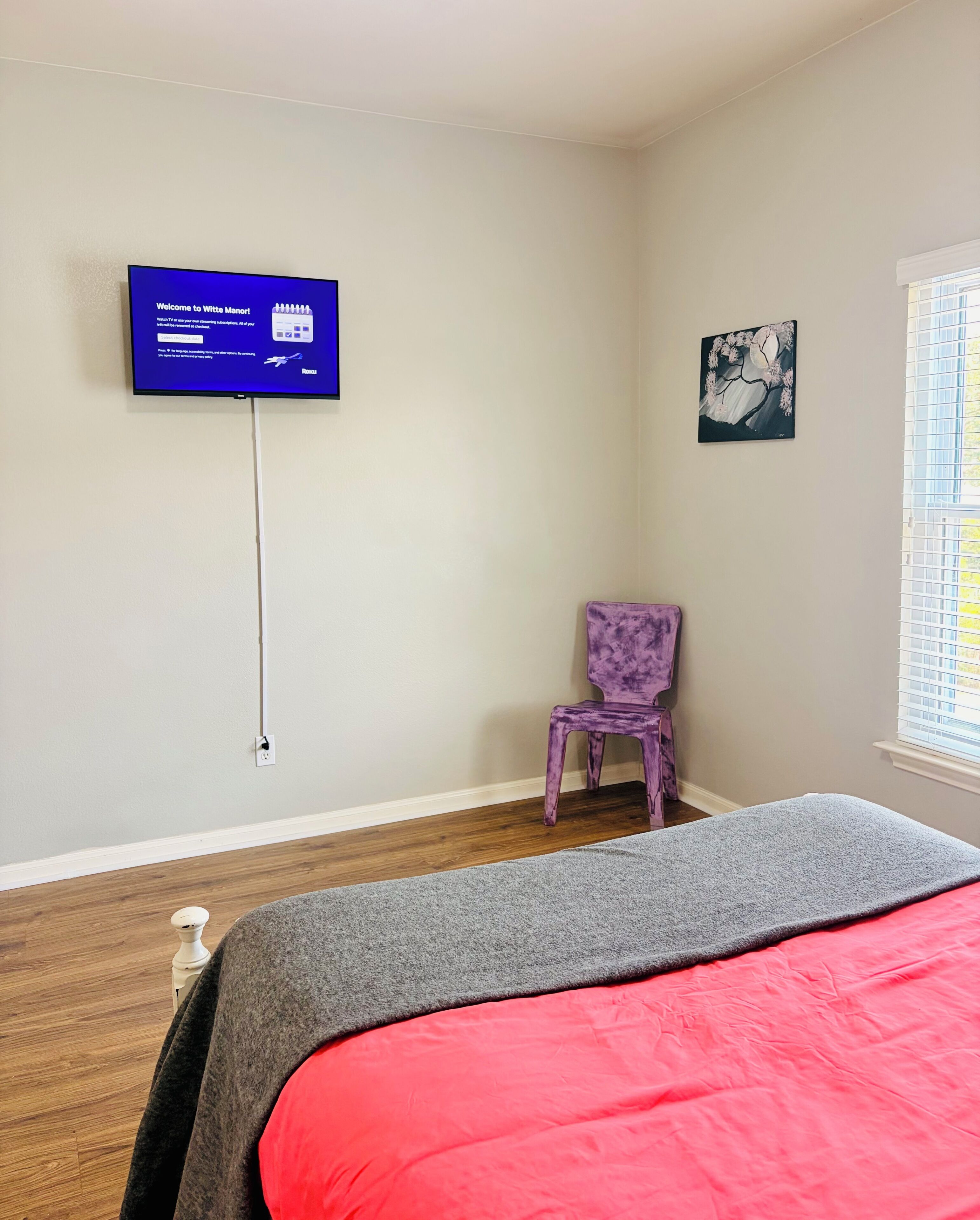 Een slaapkamer met een bed, een televisie aan de muur, een stoel en een raam met zonwering.