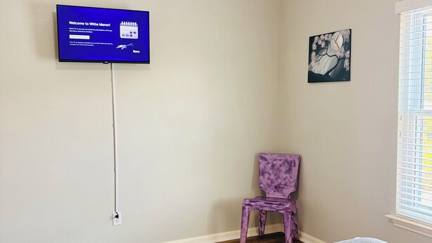 Ein Schlafzimmer mit einem Bett, einem an der Wand befestigten Fernseher, einem Sessel und einem Fenster mit Jalousien.