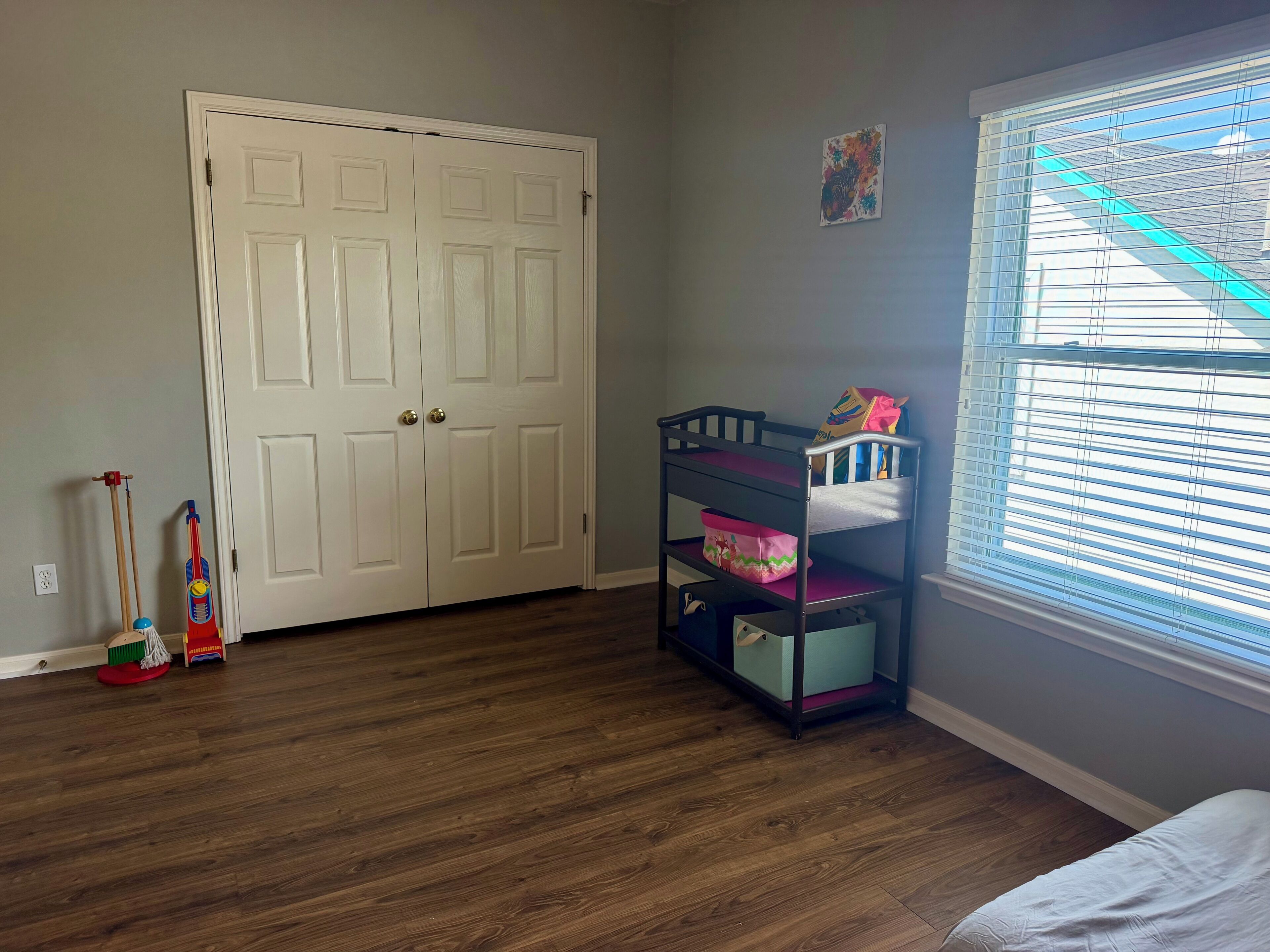 Une pièce avec un plancher en bois, une porte blanche, un lit superposé avec espace de rangement, une fenêtre avec des stores et un petit tableau accroché au mur.