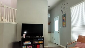Ein Wohnzimmer mit einem Flachbildfernseher, einer Bücherwand mit Snacks, einer Treppe und einer Tür zu einem weiteren Raum.