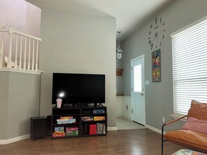 Ein Wohnzimmer mit einem Flachbildfernseher, einer Bücherwand mit Snacks, einer Treppe und einer Tür zu einem weiteren Raum.