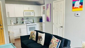 Ein kompakter Wohnbereich mit einem dunkelgrauen Sofa, einem kleinen runden Tisch und einem Deckenventilator. Im Hintergrund ist eine Küche mit weißen Schränken und einer Mikrowelle zu sehen.