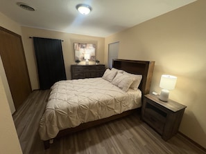 Ein Schlafzimmer mit Bett, Nachttisch, Schminktisch, einem Fenster mit Jalousien, einer Tür und einer Deckenleuchte.