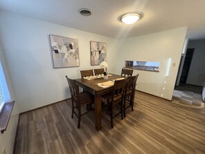 A dining area with a wooden table and chairs, two abstract paintings on the wall, and a kitchen visible through an open doorway.