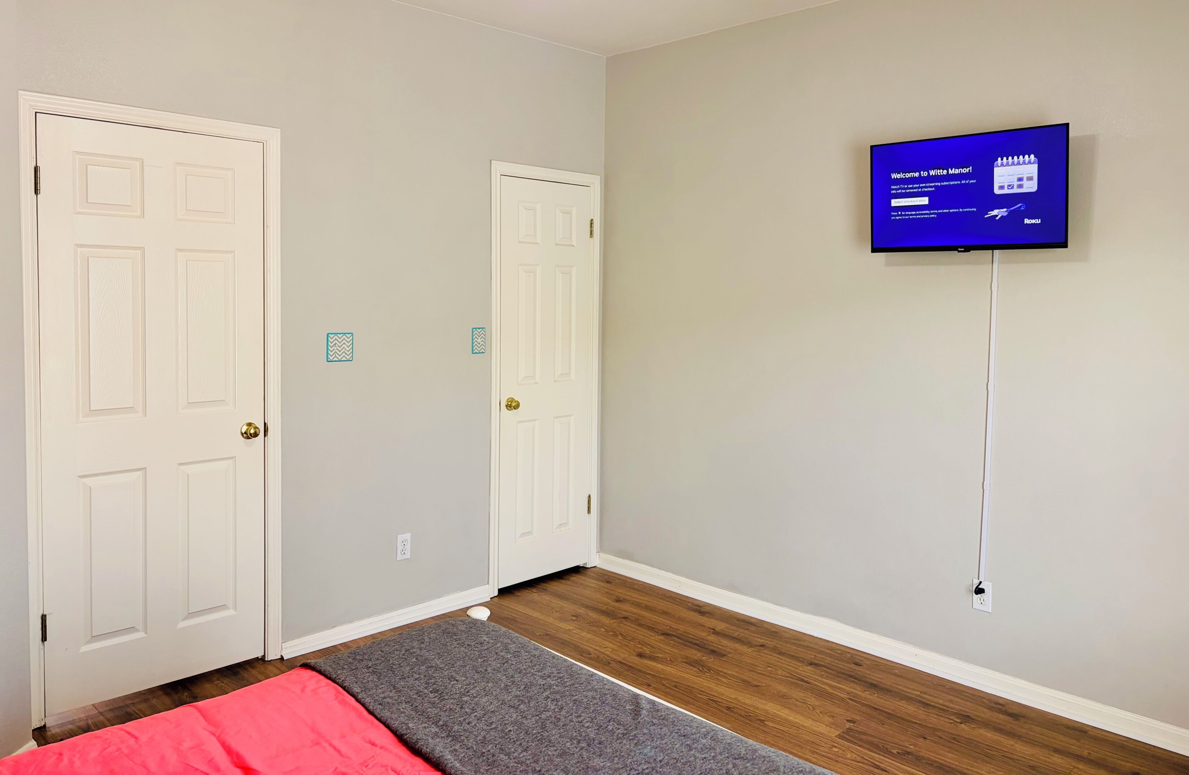 Una habitación con dos puertas blancas, un televisor de pantalla plana montado en la pared y suelo de madera.