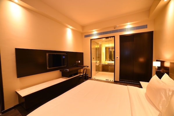 A hotel room with a bed, a TV mounted on the wall, a desk with a chair, a mirror, and a sliding door leading to a bathroom.