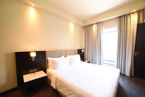 Habitación de hotel con una cama grande, mesitas de noche, un velador, una ventana con cortinas y una puerta.