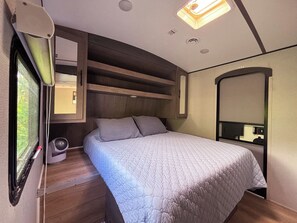A compact bedroom with a bed, a small bedside table, a fan, and a mirror.