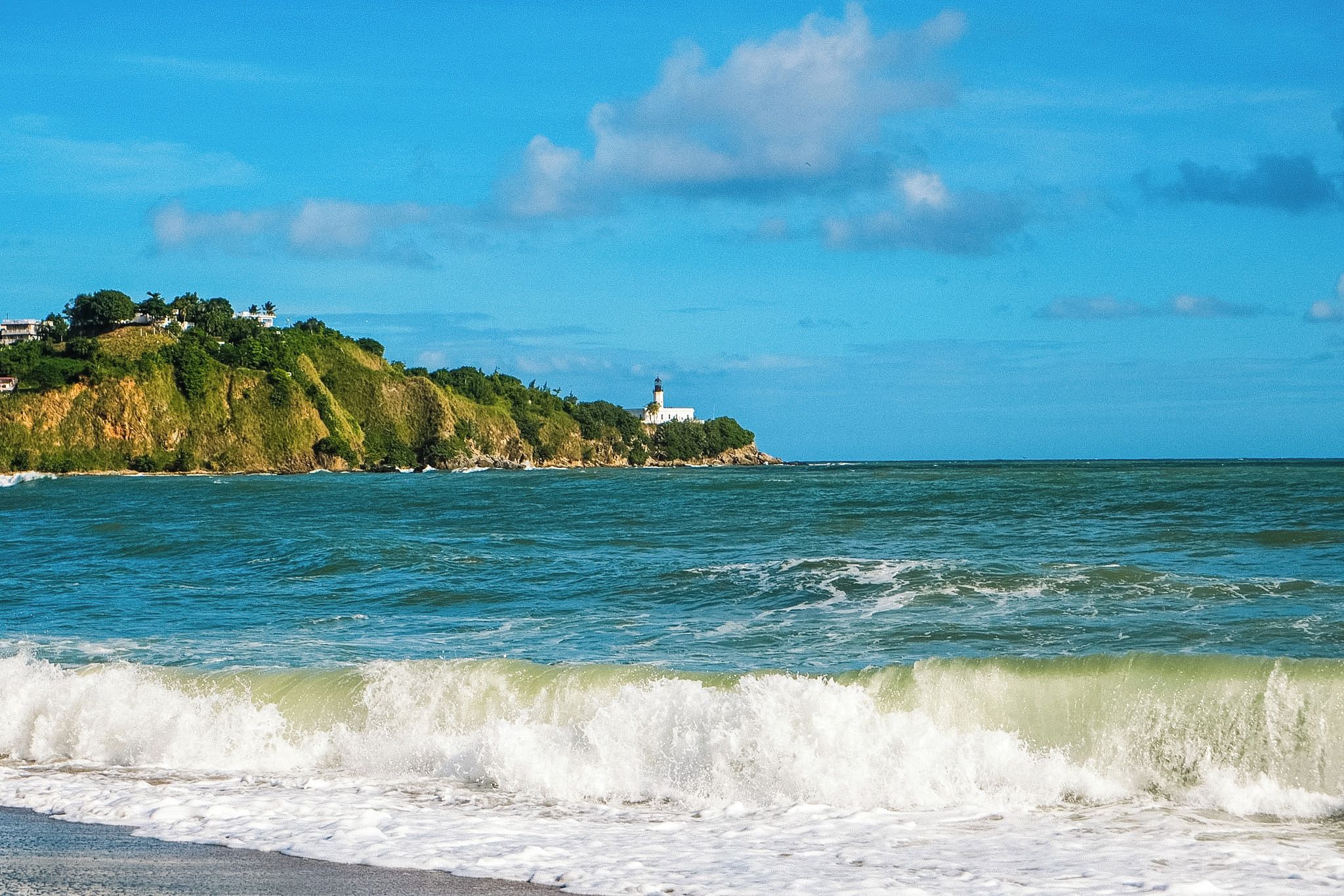 A coastal scene with a lighthouse on a hill, ocean waves, and a clear sky.