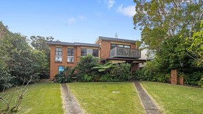 Um edifício residencial de dois andares com varanda, cercado por vegetação exuberante e um gramado bem cuidado.