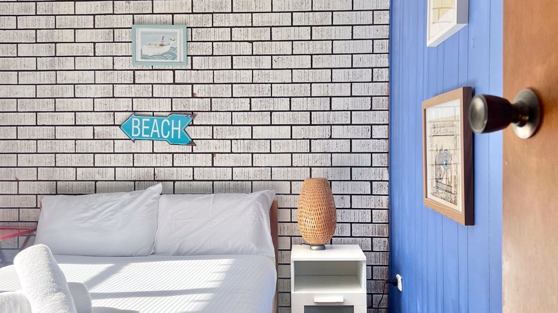 A room with a brick-patterned wall, a bed with white bedding, a small bedside table with a lamp, and a door with a blue frame.