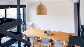 A dining area with a wooden table and white chairs, a woven pendant light, and a view through a window. - Aqua, Pet Friendly Holiday Home Close to the Beach (Manyana)