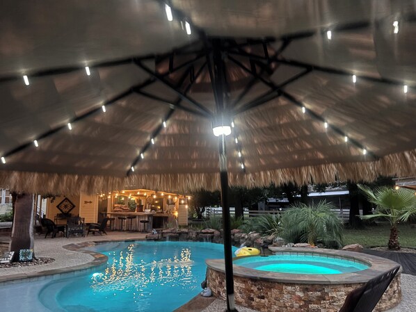 An outdoor pool area with a thatched roof and string lights, surrounded by stone walls and greenery.